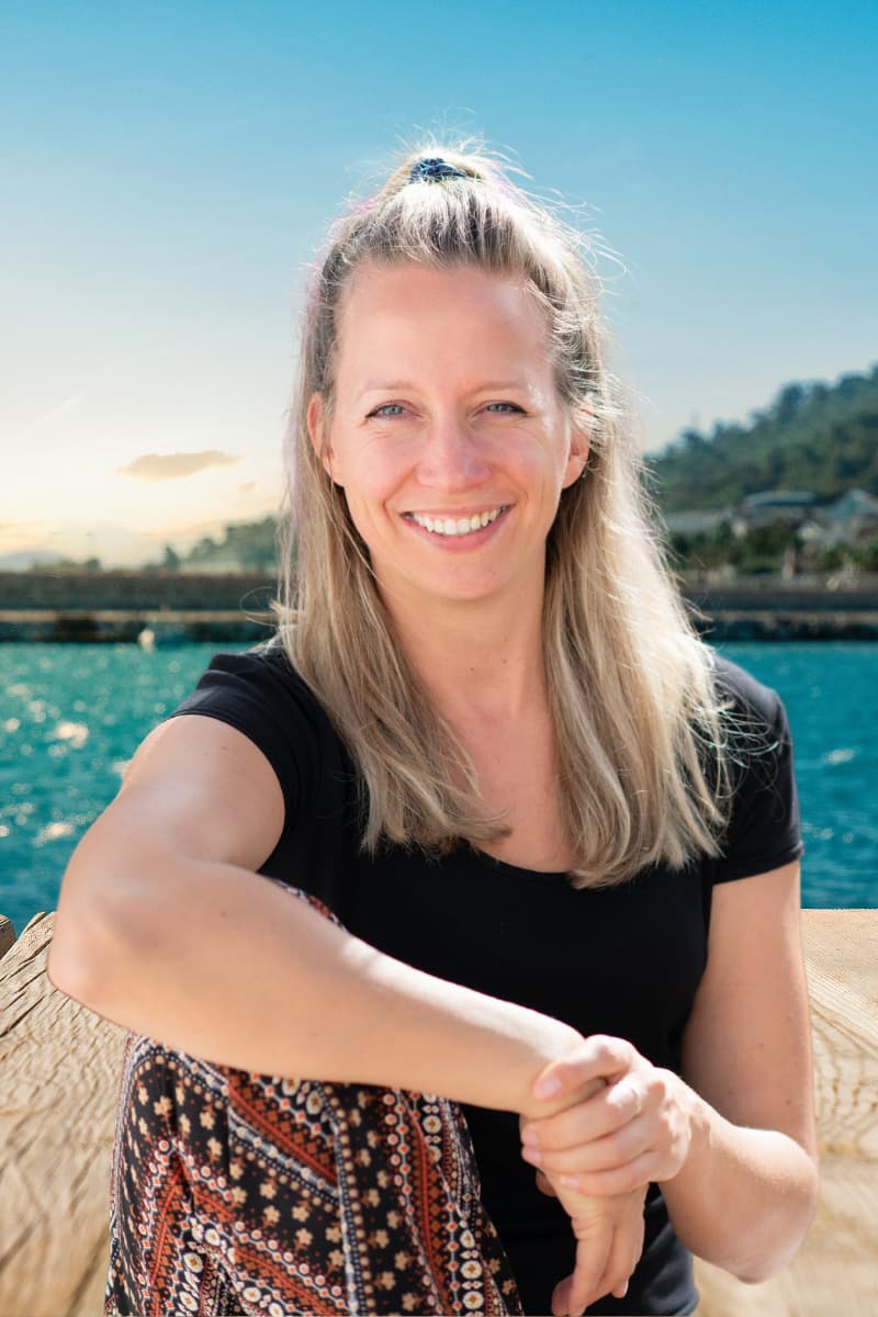 Vrouwelijke Hashimoto expert en trage schildklier specialist zit lachend aan het water; holistische focus op voeding, dieet, afvallen, meer energie en gezondheid.