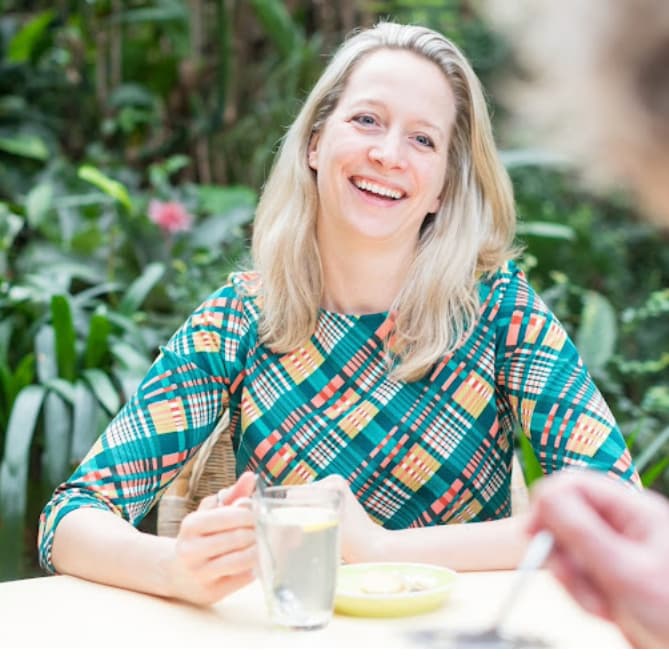 Marloes Mulder die ontspannen aan tafel zit met een kop thee, in een rustige groene omgeving tijdens een warm gesprek over gezondheid en herstel.