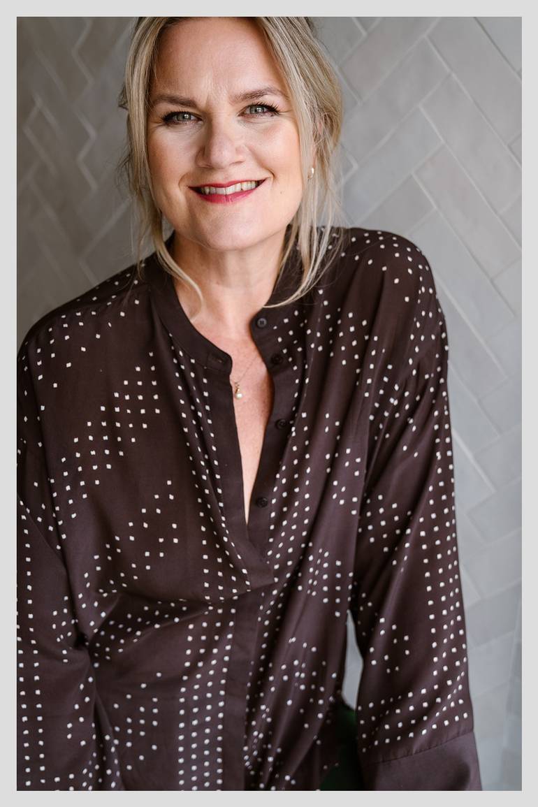 Ninke van der Leck glimlacht zelfverzekerd in een donkerbruin blouse met witte stippen, zittend voor een lichte tegelwand. Dankzij begeleiding bij Hashimoto zegt ze: "Mijn schildklier ging beter functioneren — iets waarvan artsen zeiden dat het niet kon."
